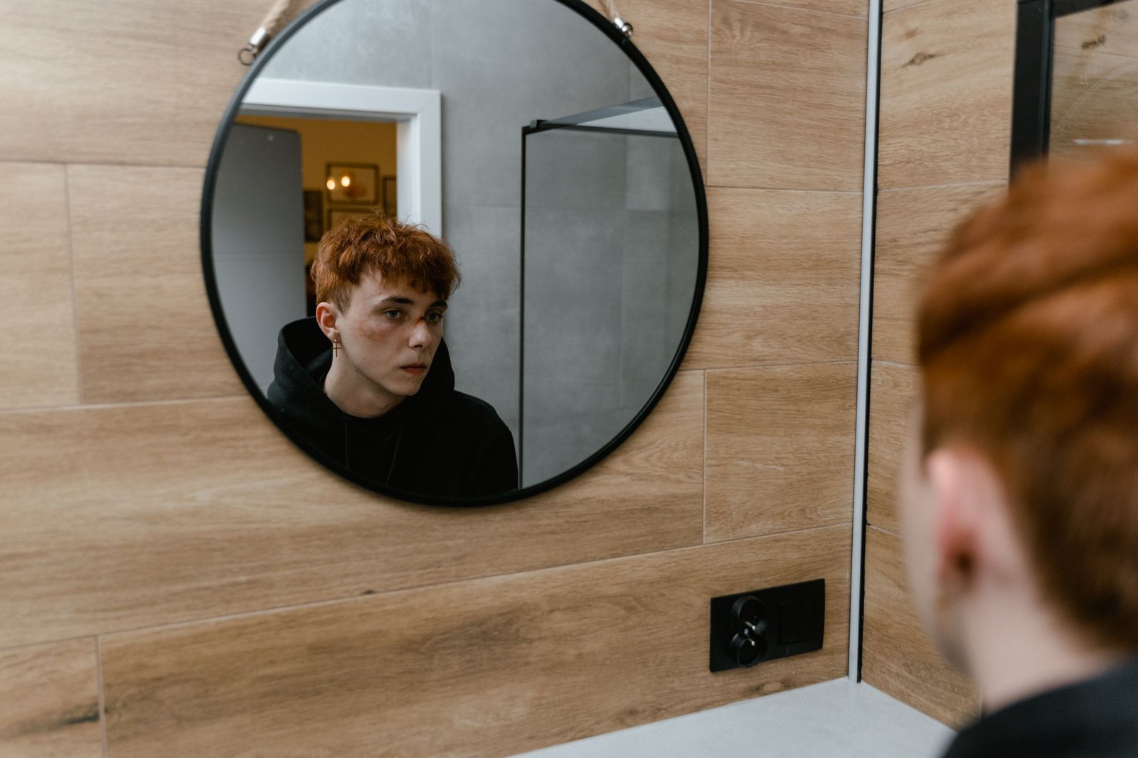 A redheaded teen boy gazes introspectively into a round mirror, reflecting a thoughtful moment.