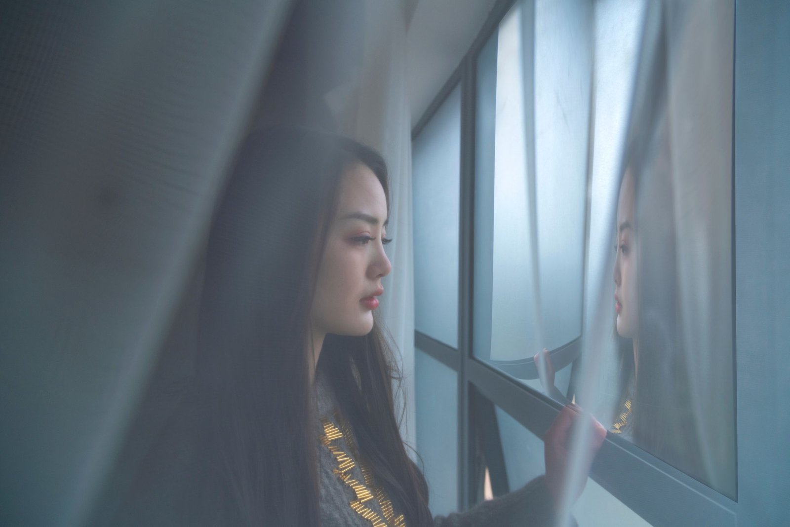 A woman stands by a window, reflecting in thought amidst soft lighting.