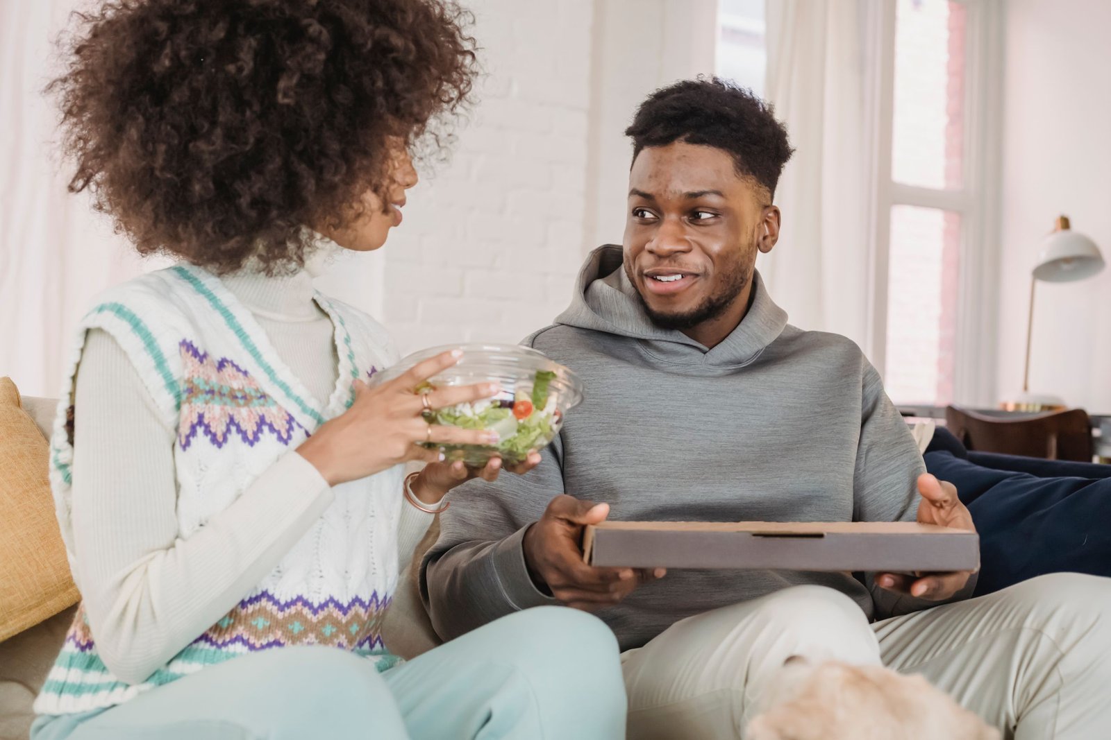 Crop young African American woman in casual clothes with curly hair eating appetizing salad while spending time together with smiling boyfriend and dog