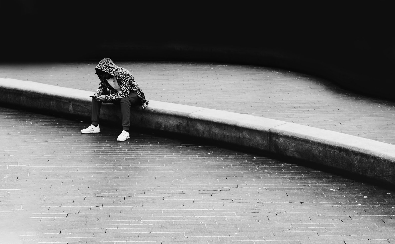 Person in a hoodie sitting on a curb in a monochrome urban setting.