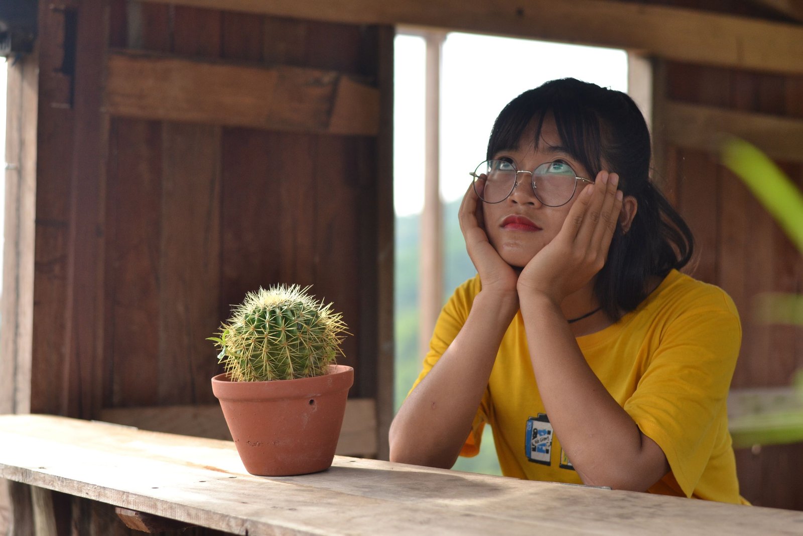 Woman in glasses and yellow shirt daydreaming beside potted cactus on a wooden table indoors.