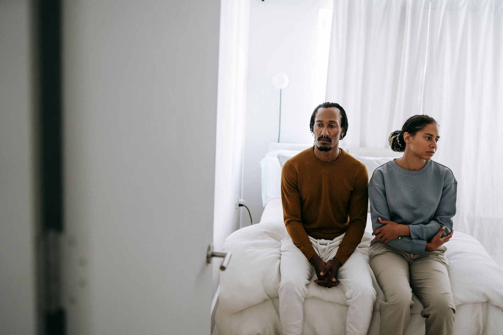 Young African American couple in casual outfit sitting on bed together with sad face in daytime